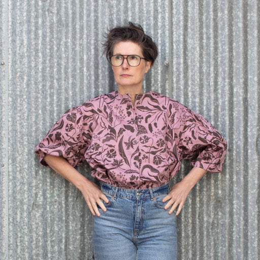 Woman wearing a blouse printed with graphic chocolate and rose coloured wildflowers. Blouse is collarless and button down with a dropped shoulder and a gathered puff sleeve